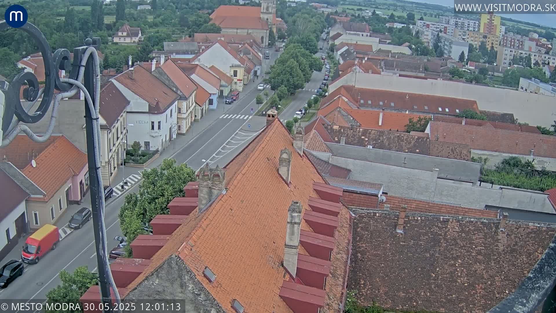 An aerial view shows a town with red-tiled rooftops and a street lined with cars under an overcast sky.