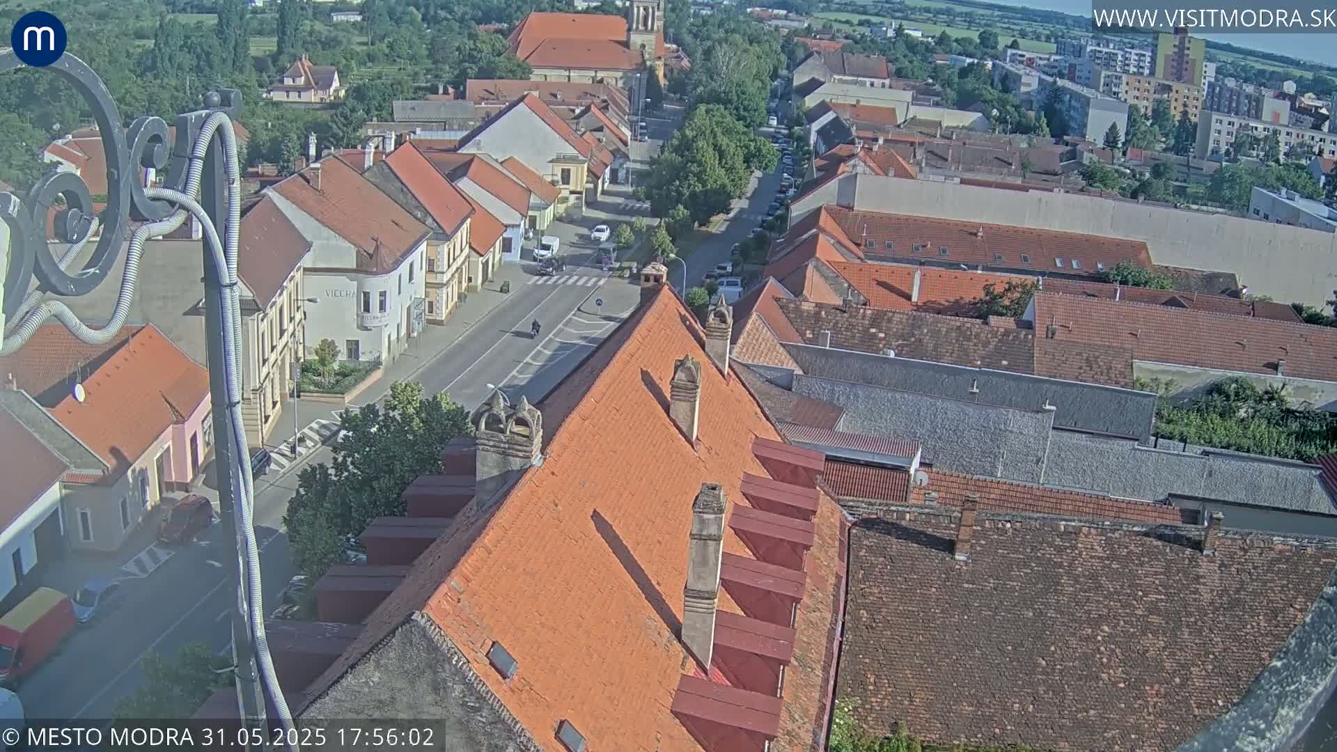 A high-angle view of a town on a sunny day, showing many red-tiled roofs and a street with sparse traffic.