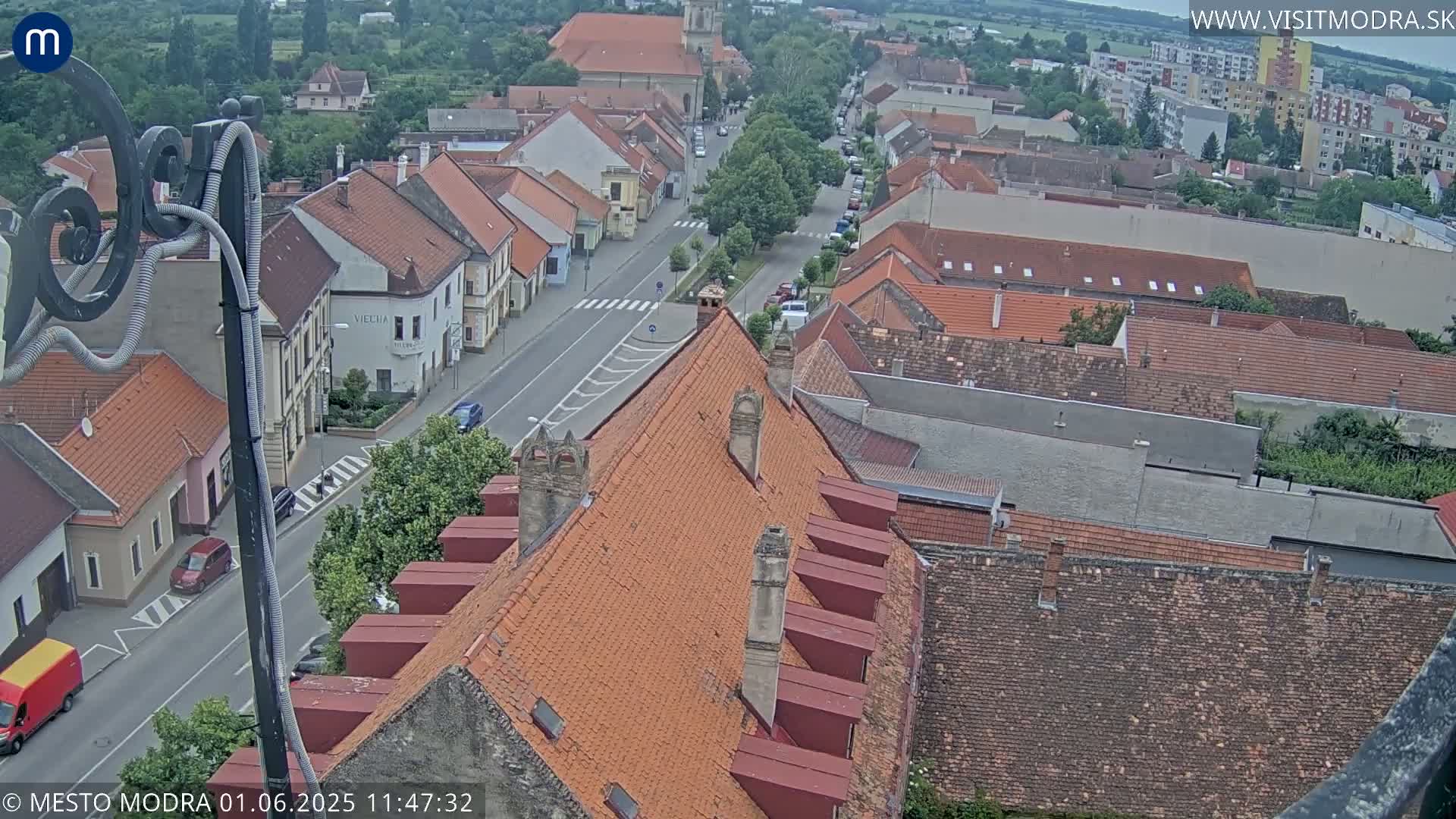 An elevated view shows a town's street and many buildings with orange-tiled roofs, under an overcast sky.