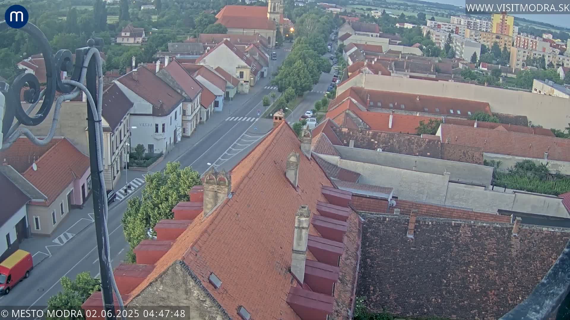An elevated view of a town with red-tiled roofs and a street running through it, under a clear, light sky.