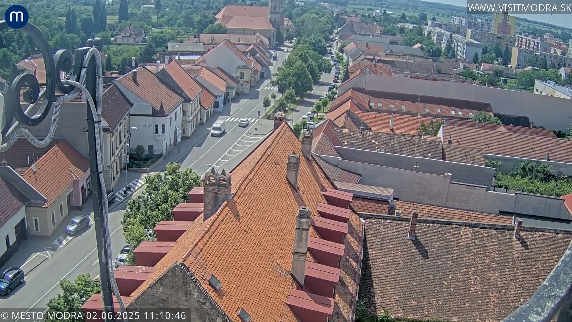 A high-angle view of a town on a sunny day, showing red-tiled rooftops, a street with cars, and surrounding buildings.