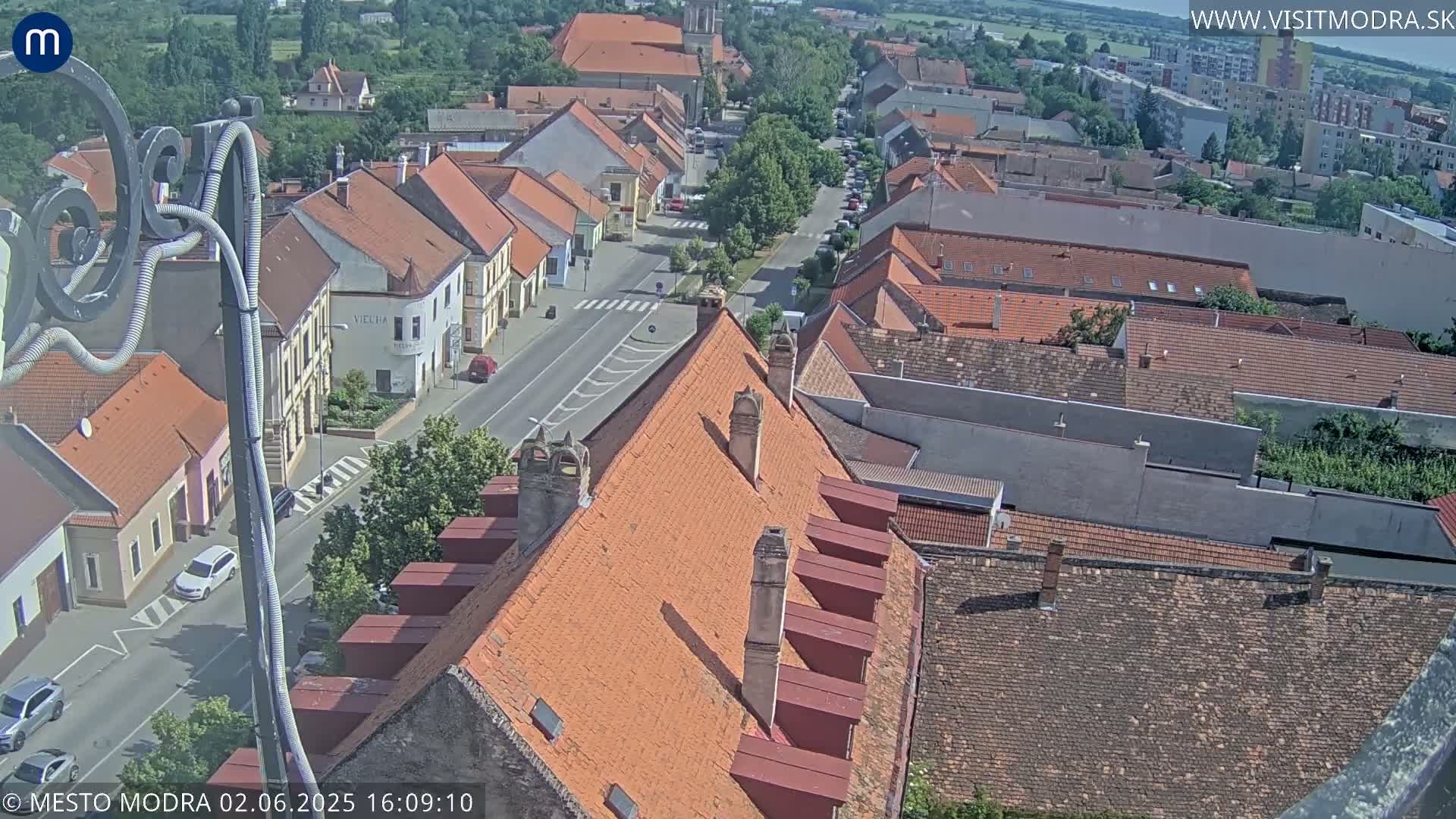 An elevated view of a town on a sunny day, showing red-tiled rooftops and a street with cars, extending into a larger landscape of buildings and greenery.