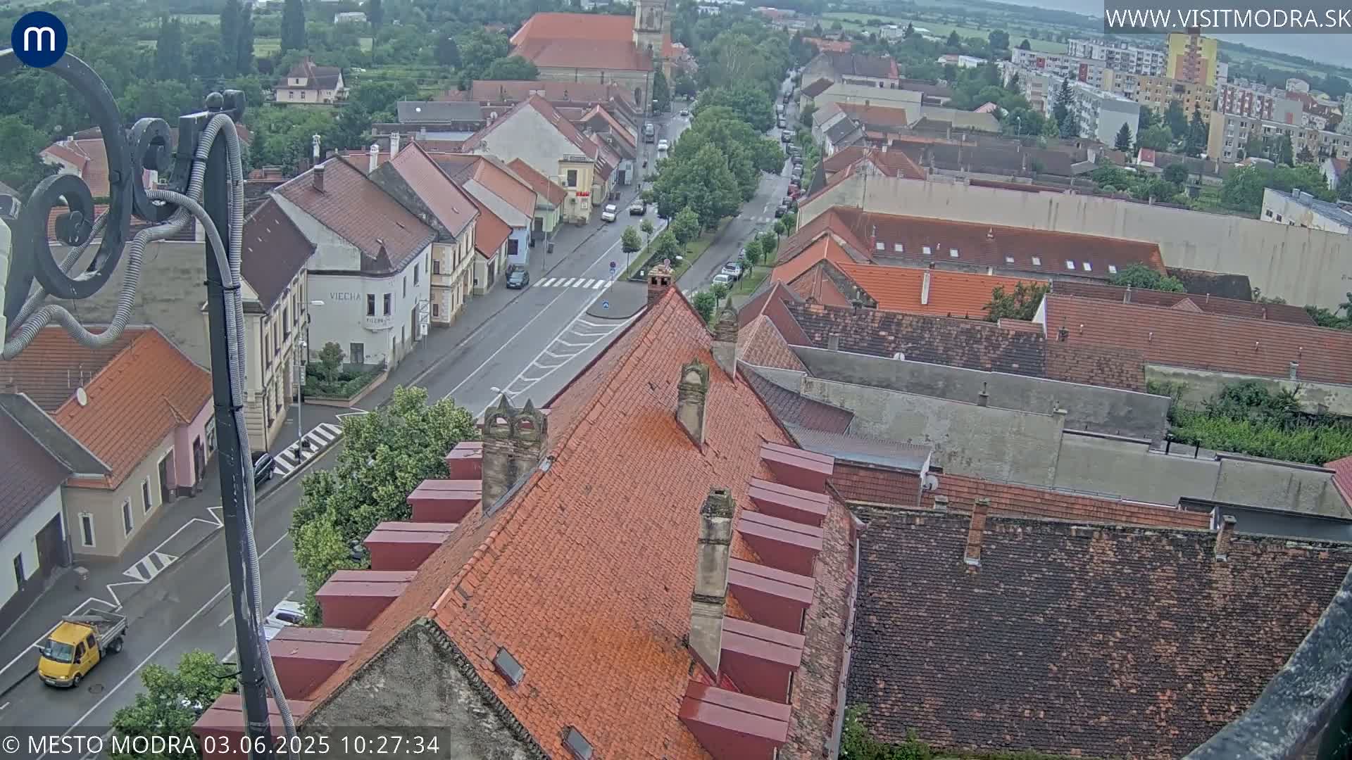 An overcast day reveals a high-angle view of a town's red-tiled rooftops and streets, with a few cars visible.