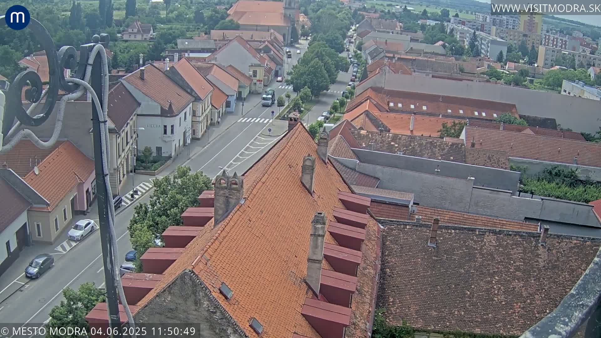 An aerial view shows a town on a partly cloudy day with red-tiled roofs and a street running through it.