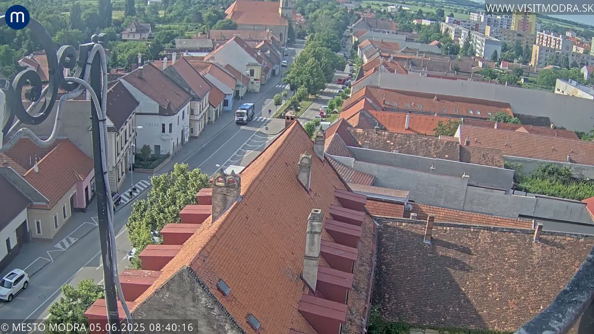 An aerial view of a town on a sunny day shows red-tiled rooftops, a street with a few vehicles, and surrounding greenery.