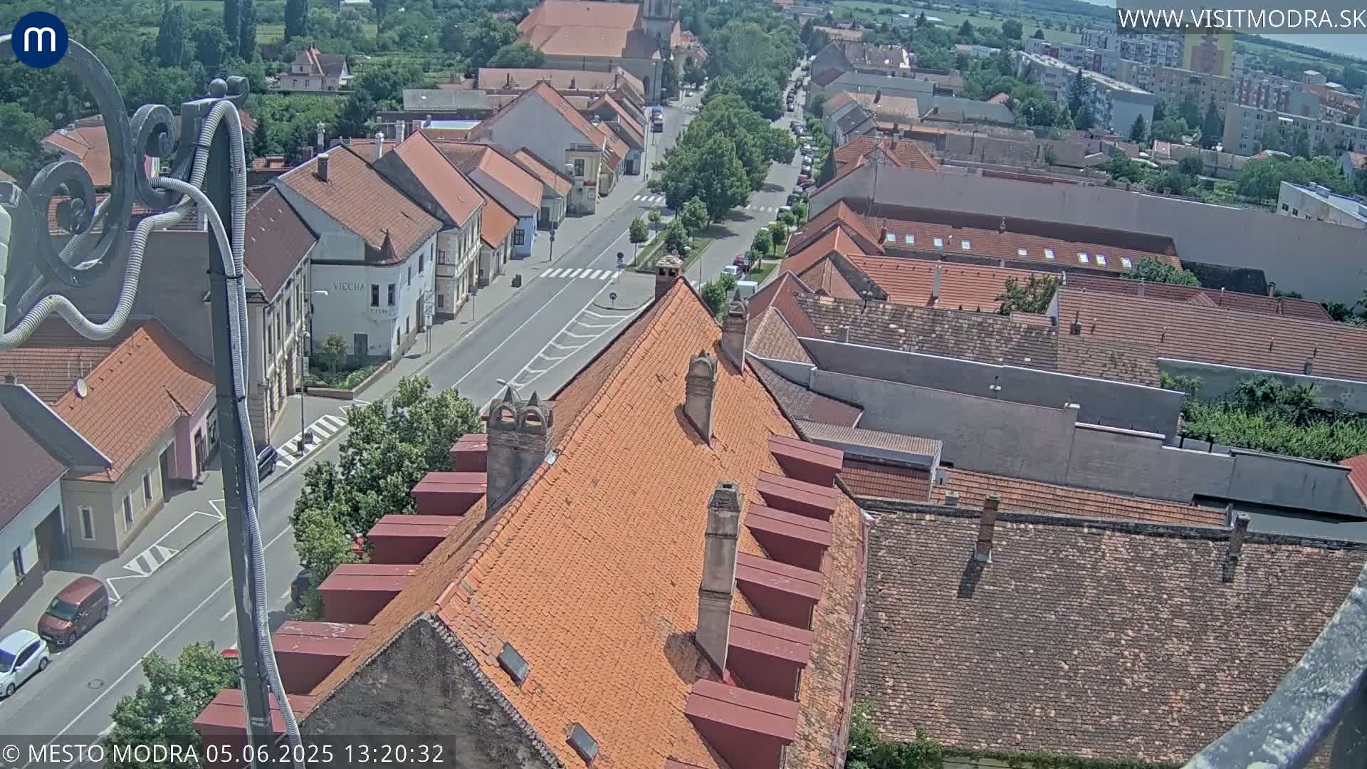 An aerial view shows a town with red-tiled roofs and a street running through it on a partly sunny day.