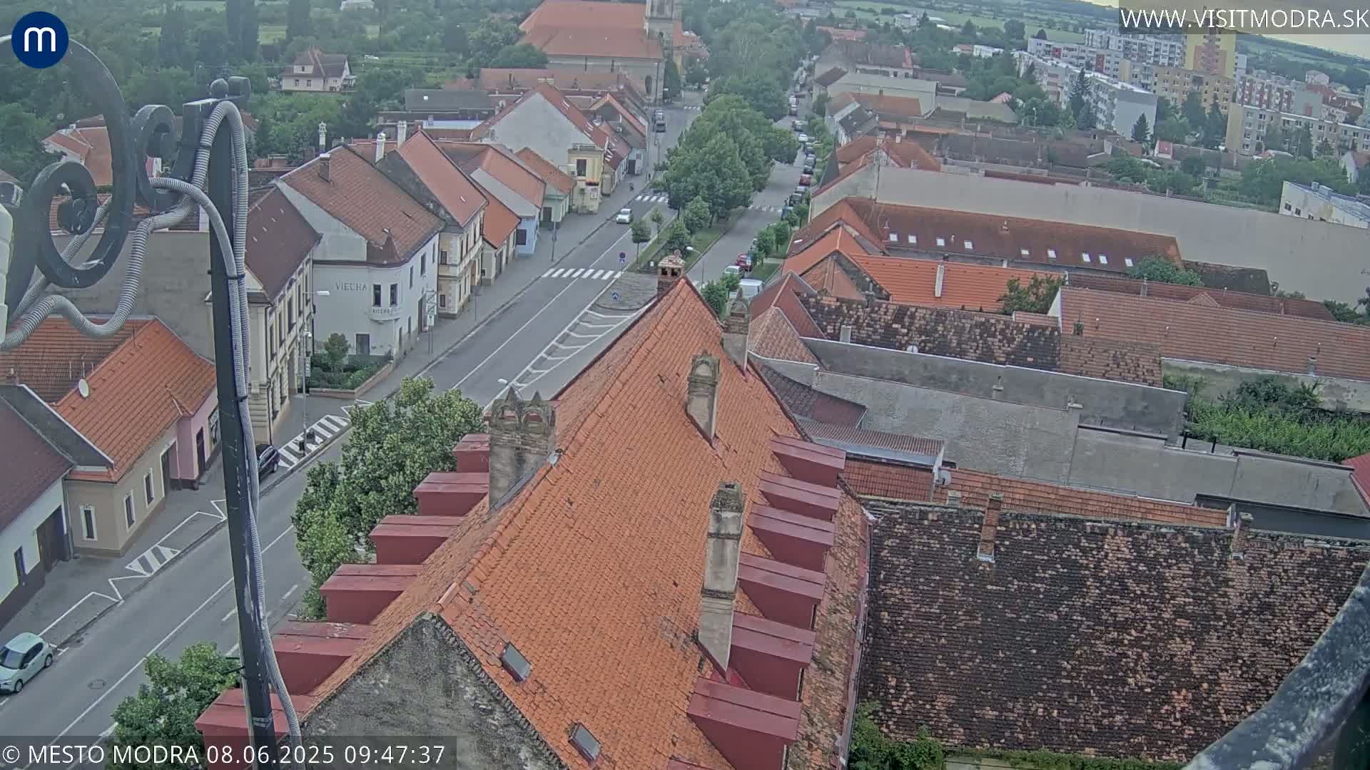 An elevated view of a town shows a street with cars and buildings with red tile roofs under an overcast sky.