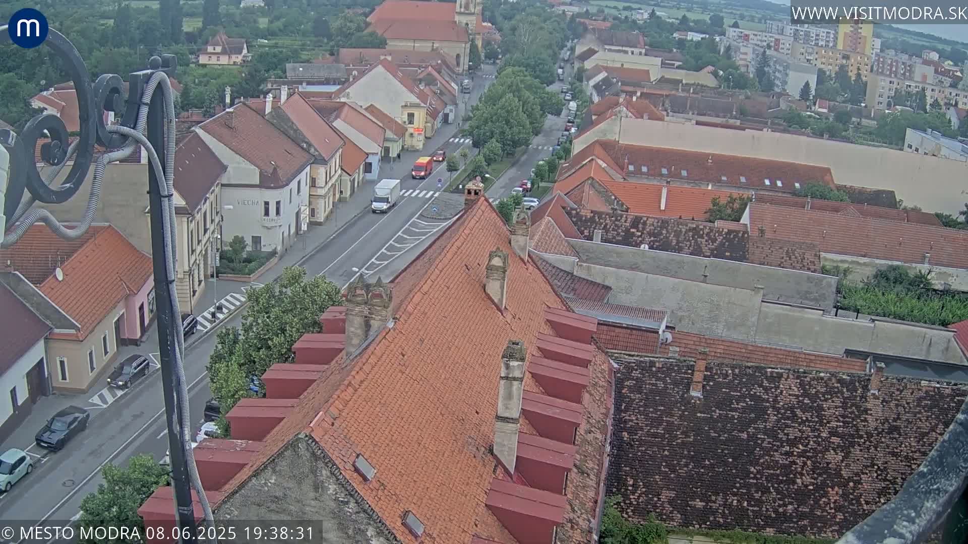 An elevated view of a town with red-tiled roofs and a street with cars, under a mostly cloudy sky.