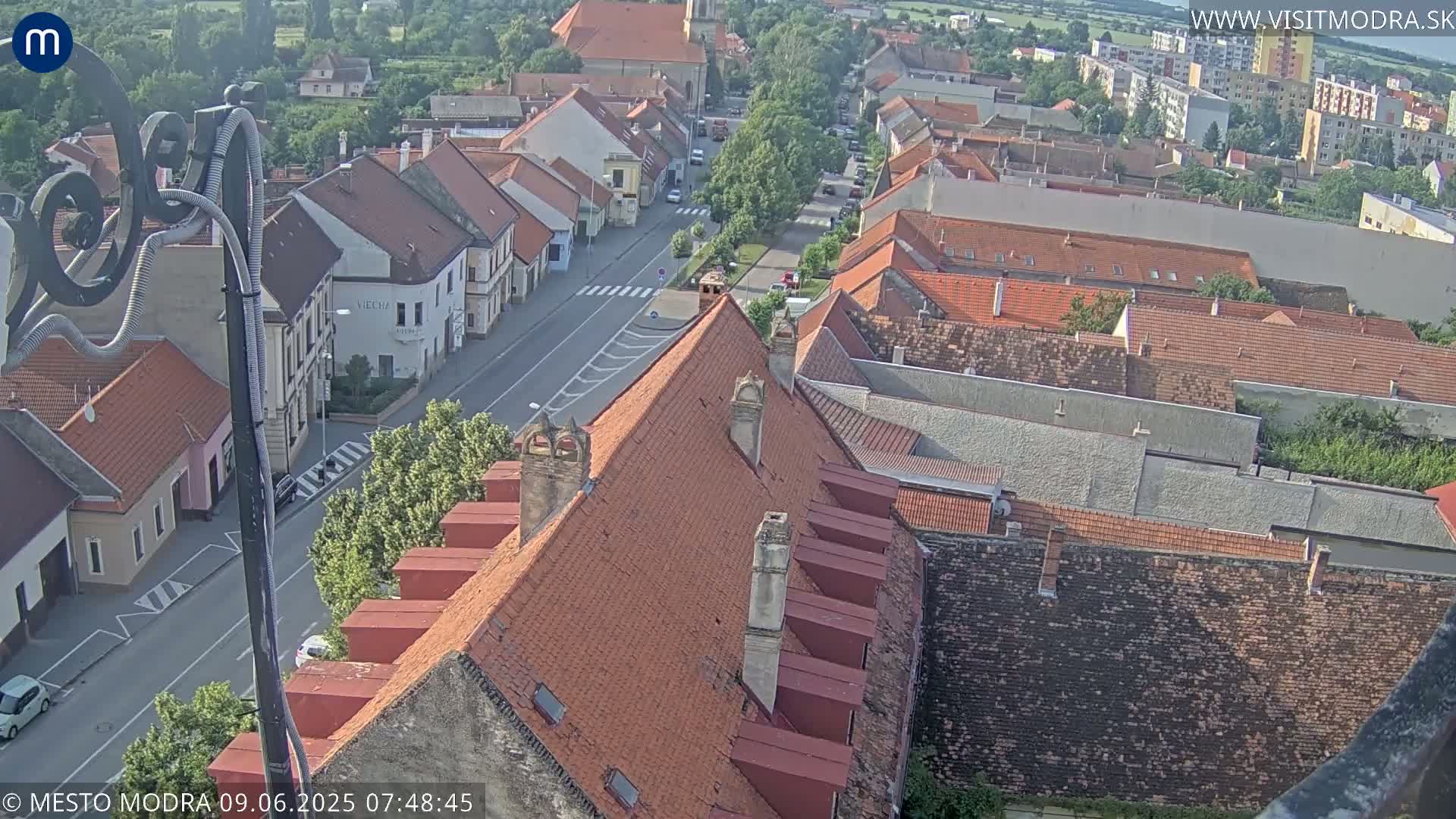 An aerial view of a town on a sunny day shows a street lined with buildings, with red-tiled rooftops dominating the scene.