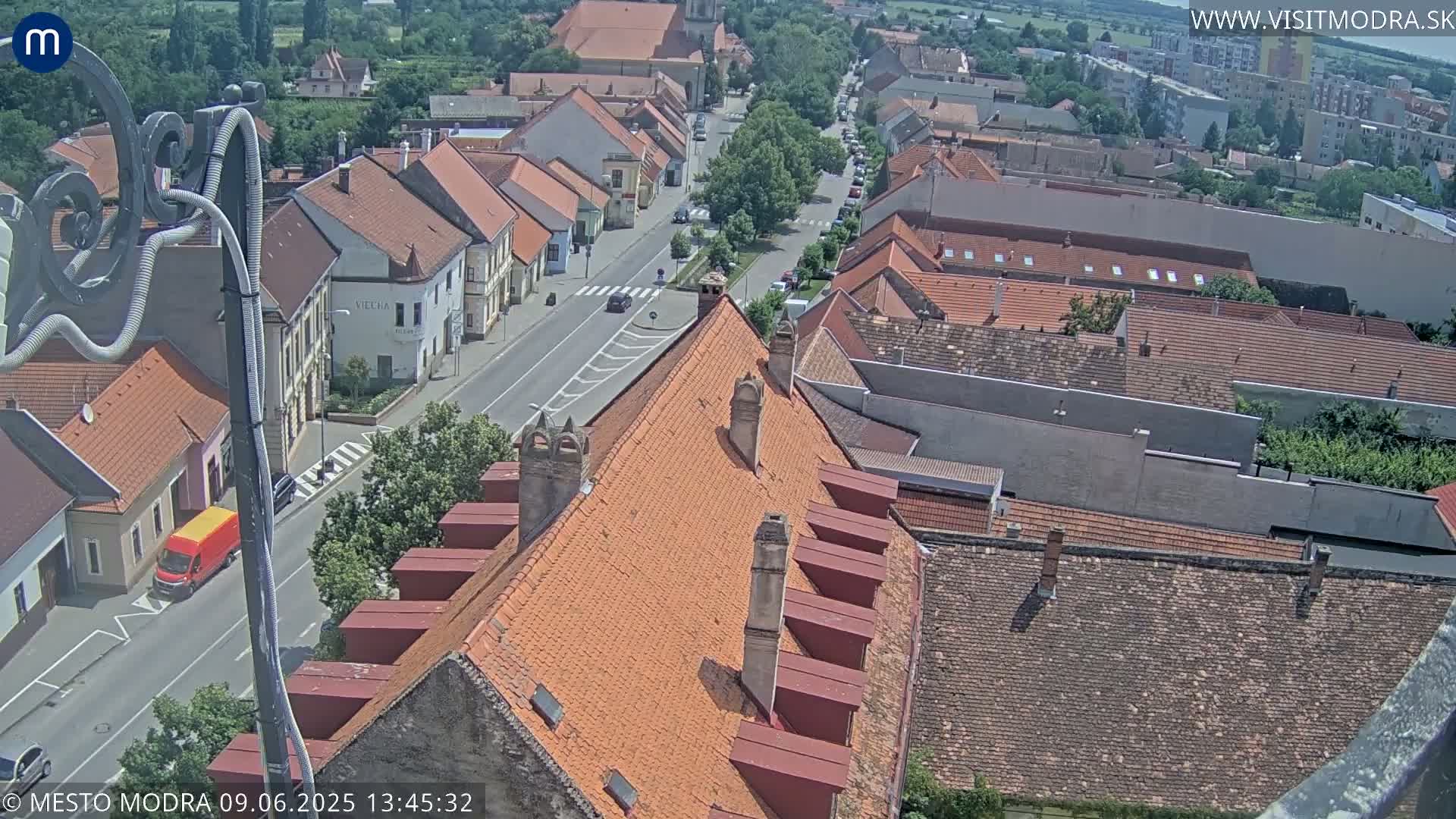 An aerial view of a town on a sunny day, showing mostly red-tiled roofs and a street with cars.