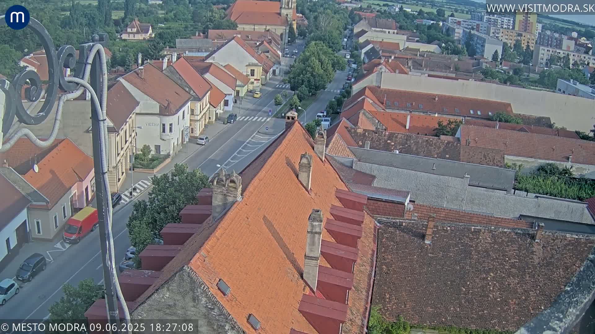 An elevated view of a town on a sunny day, showing red-tiled rooftops and a street with parked cars.