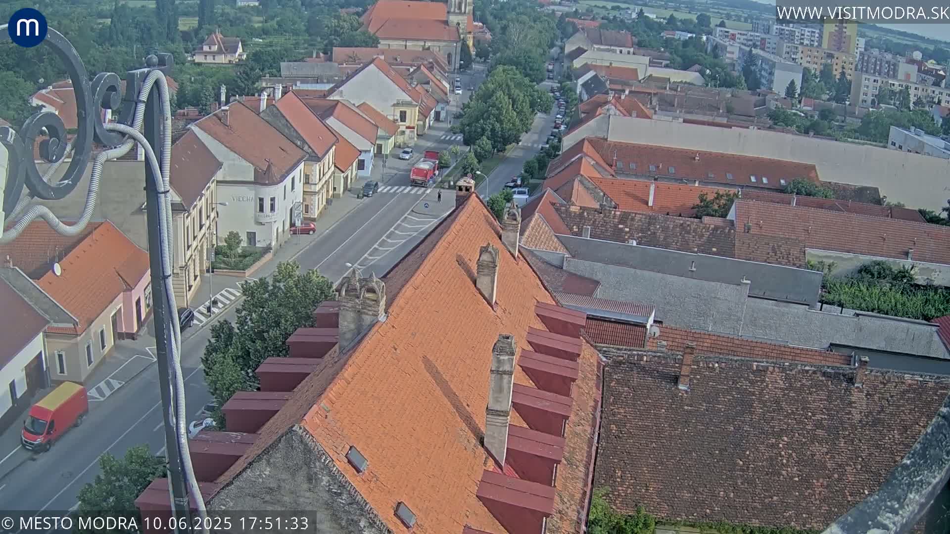 An aerial view of a town on a sunny day shows a street with cars and buildings with red-tiled roofs.