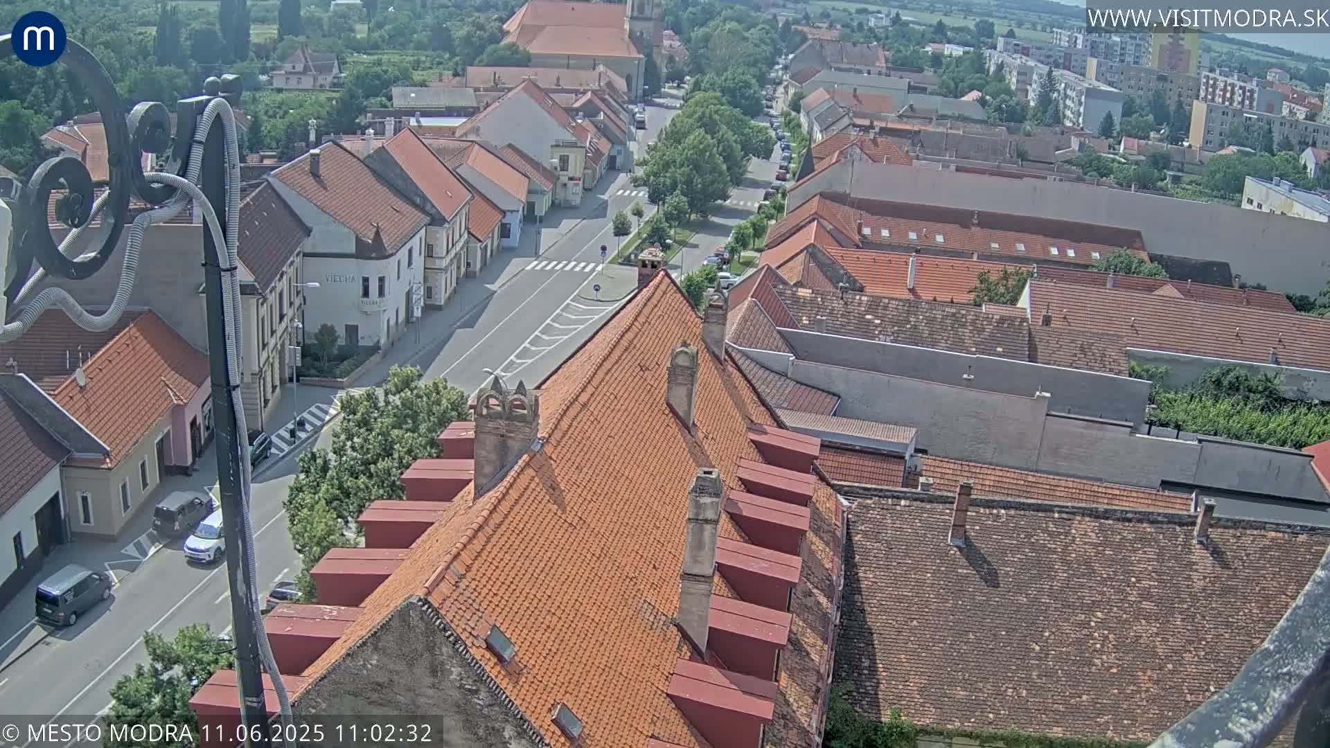 A high-angle, sunny view of a town's red-tiled rooftops and streets, with cars parked along the roads.