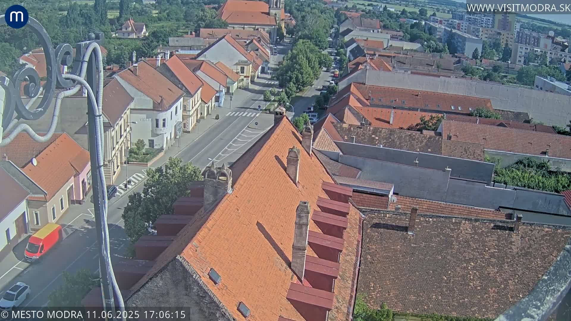 An elevated view of a town on a sunny day shows a network of red-tiled roofs, narrow streets, and sparse vehicle traffic.