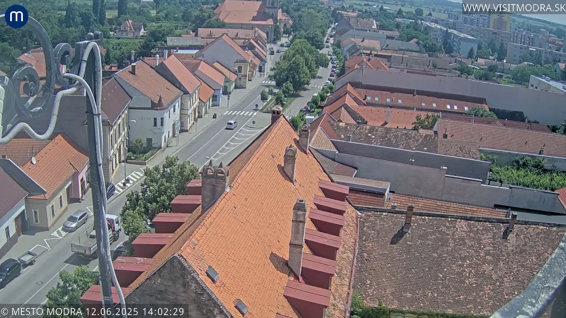An elevated view of a town on a sunny day shows a street lined with red-tiled buildings, with several cars visible on the road.