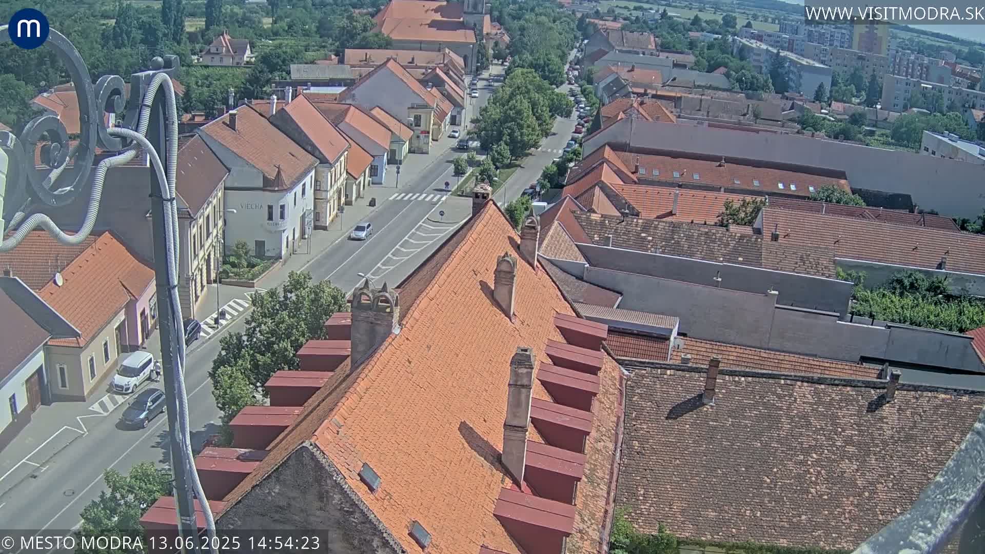 An aerial view of a town with red-tiled roofs on a sunny day, showing a street with cars and surrounding buildings.
