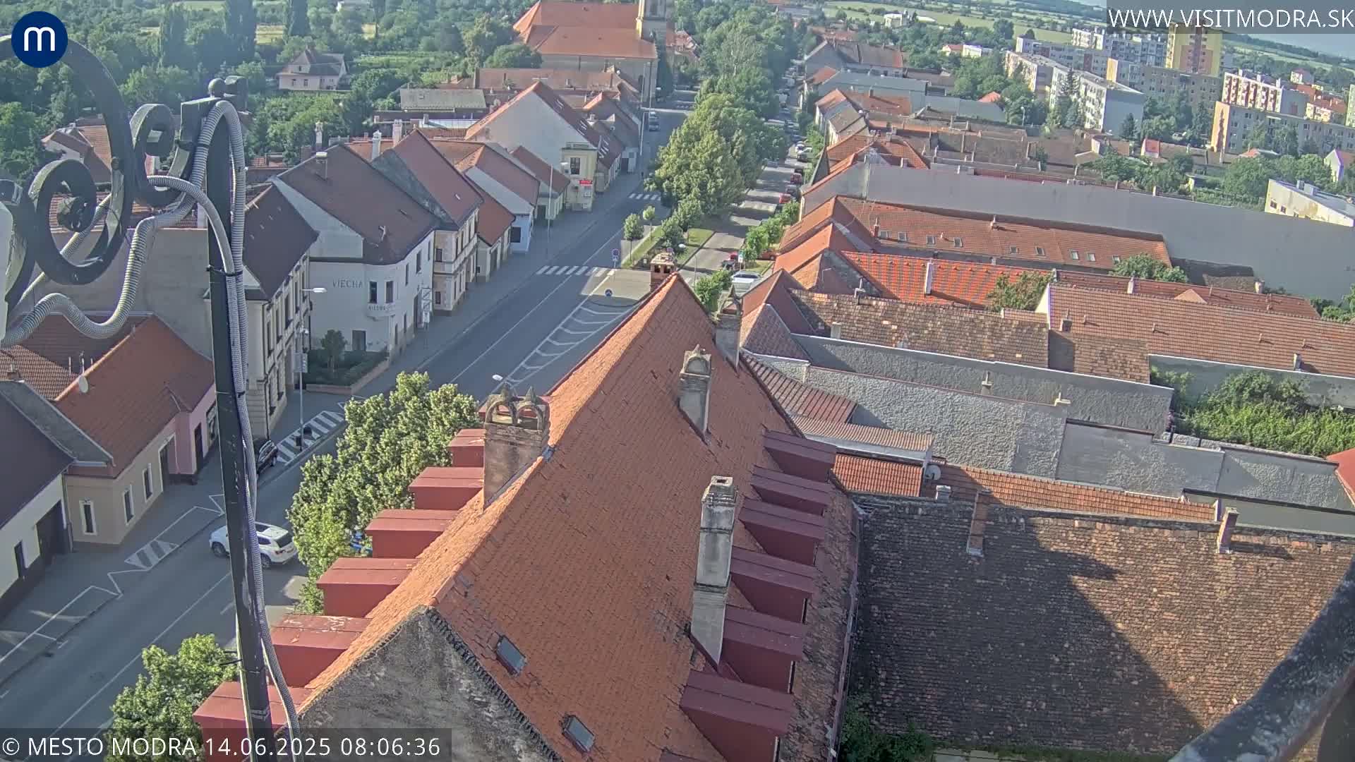 An aerial view shows a town with red-tiled roofs and a street below, on a sunny day.
