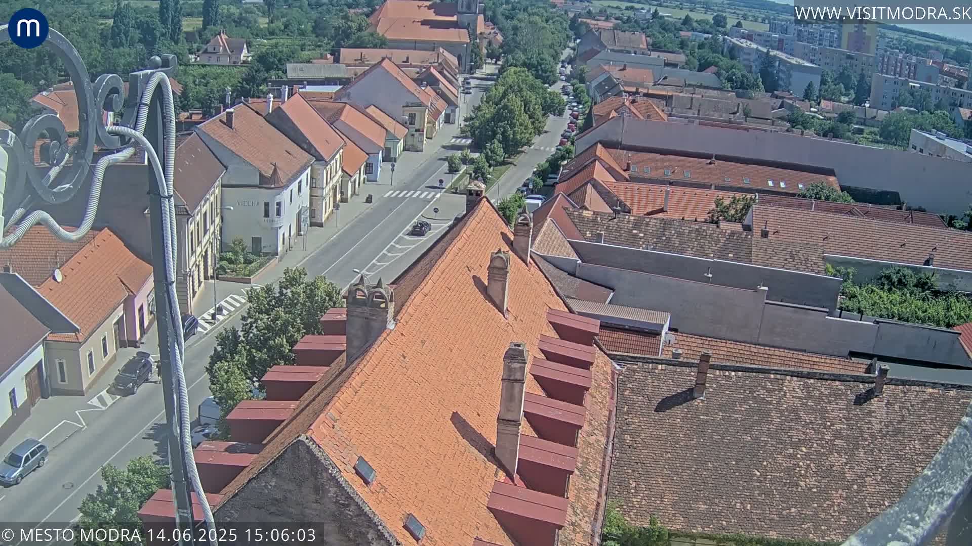 An elevated view of a town on a sunny day, showing red-tiled rooftops, narrow streets, and a few cars.