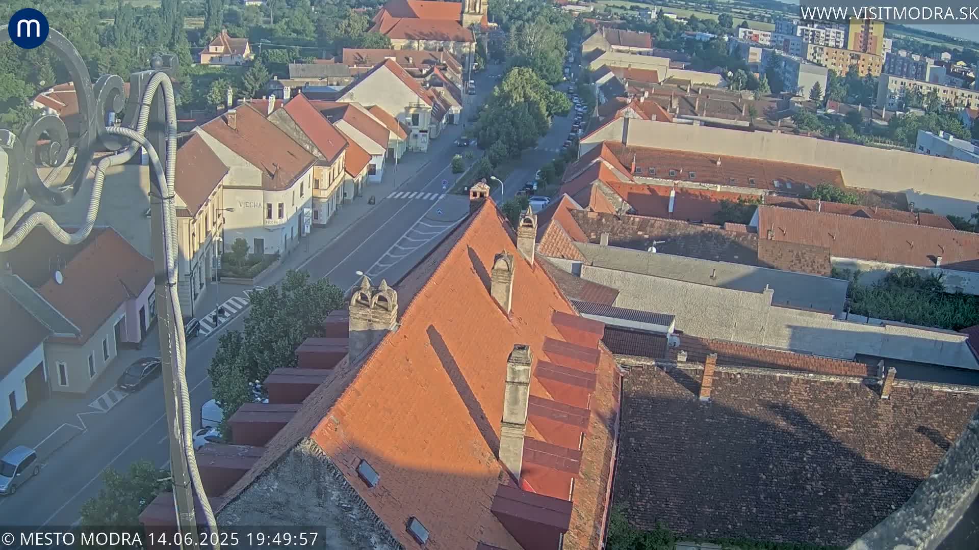 An aerial view shows a town with red-tiled roofs under a clear, sunny sky.