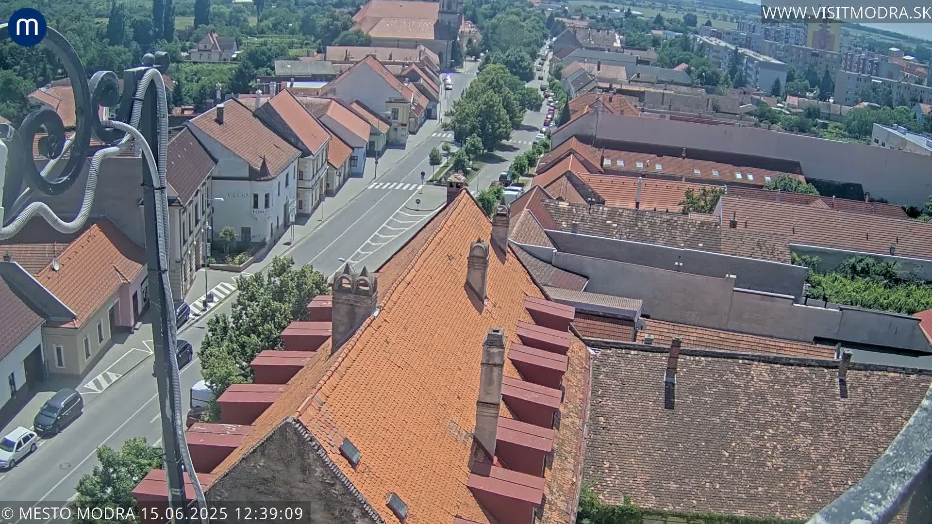 An aerial view of a town on a sunny day, showing a street lined with red-tiled buildings, viewed from a high vantage point overlooking a section of orange tiled rooftops.