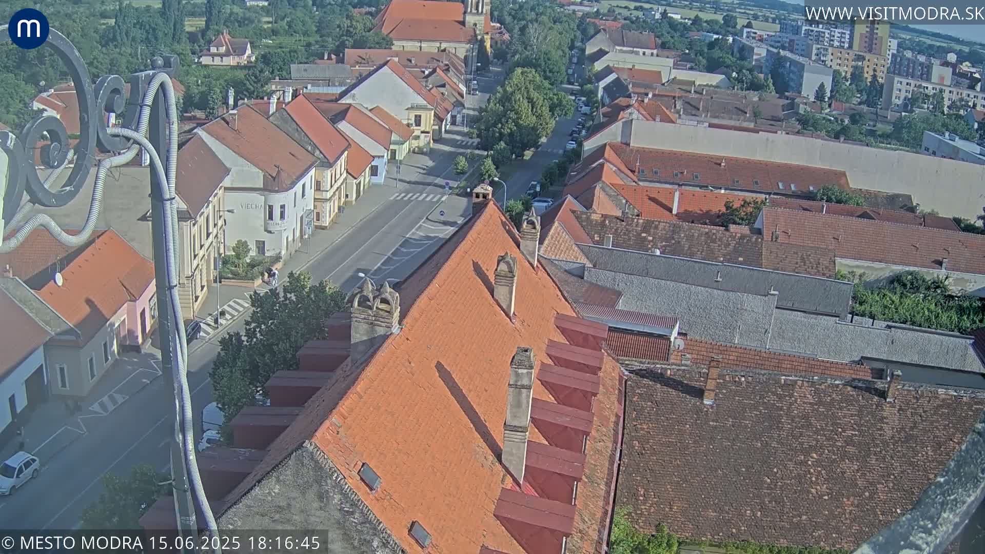 An aerial view of a town on a sunny day shows numerous red-tiled roofs, a street with cars, and a section of a larger building with an orange roof and multiple chimneys.