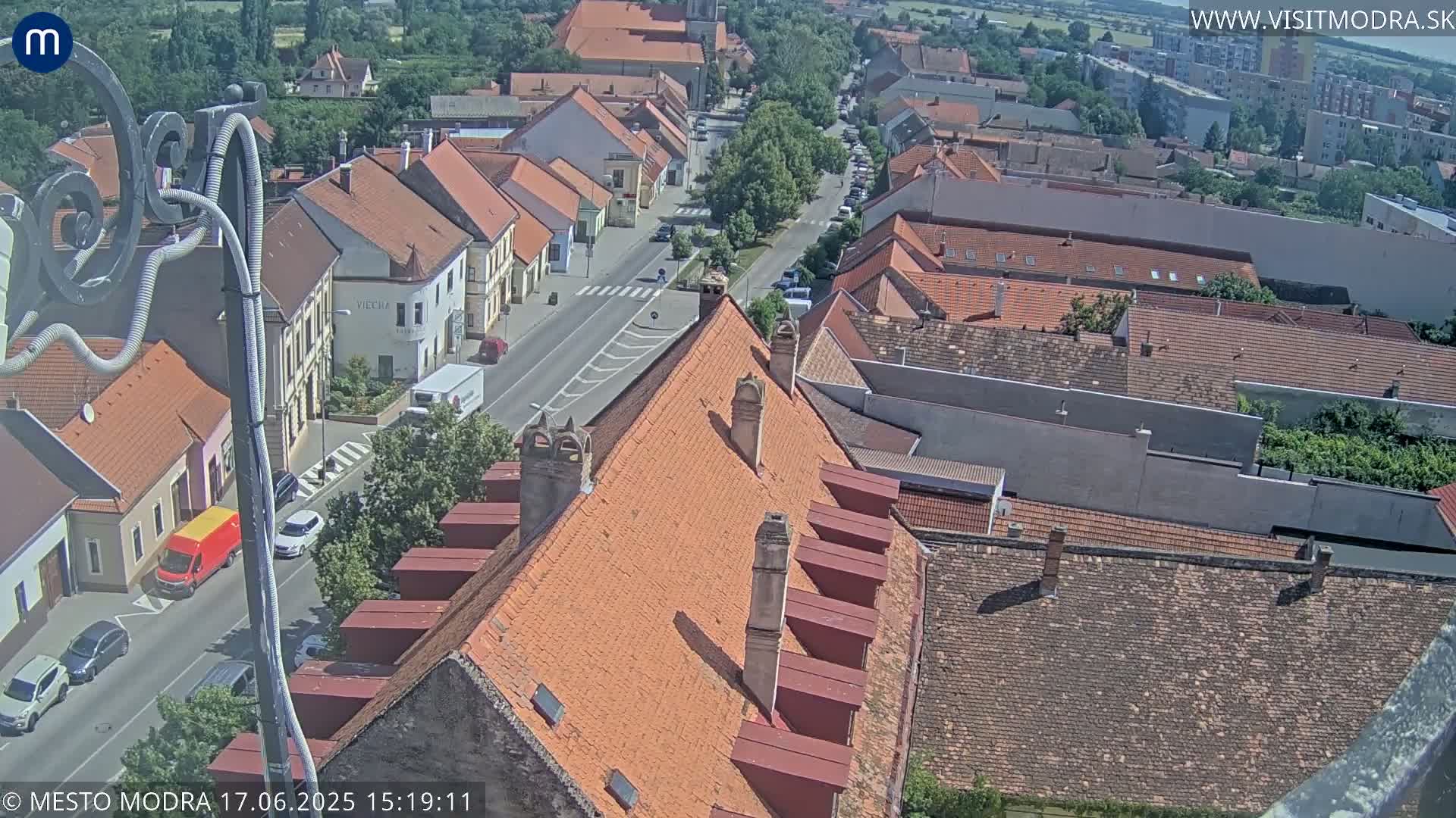A high-angle view on a sunny day shows a town's rooftops and a street with cars, extending into the distance.