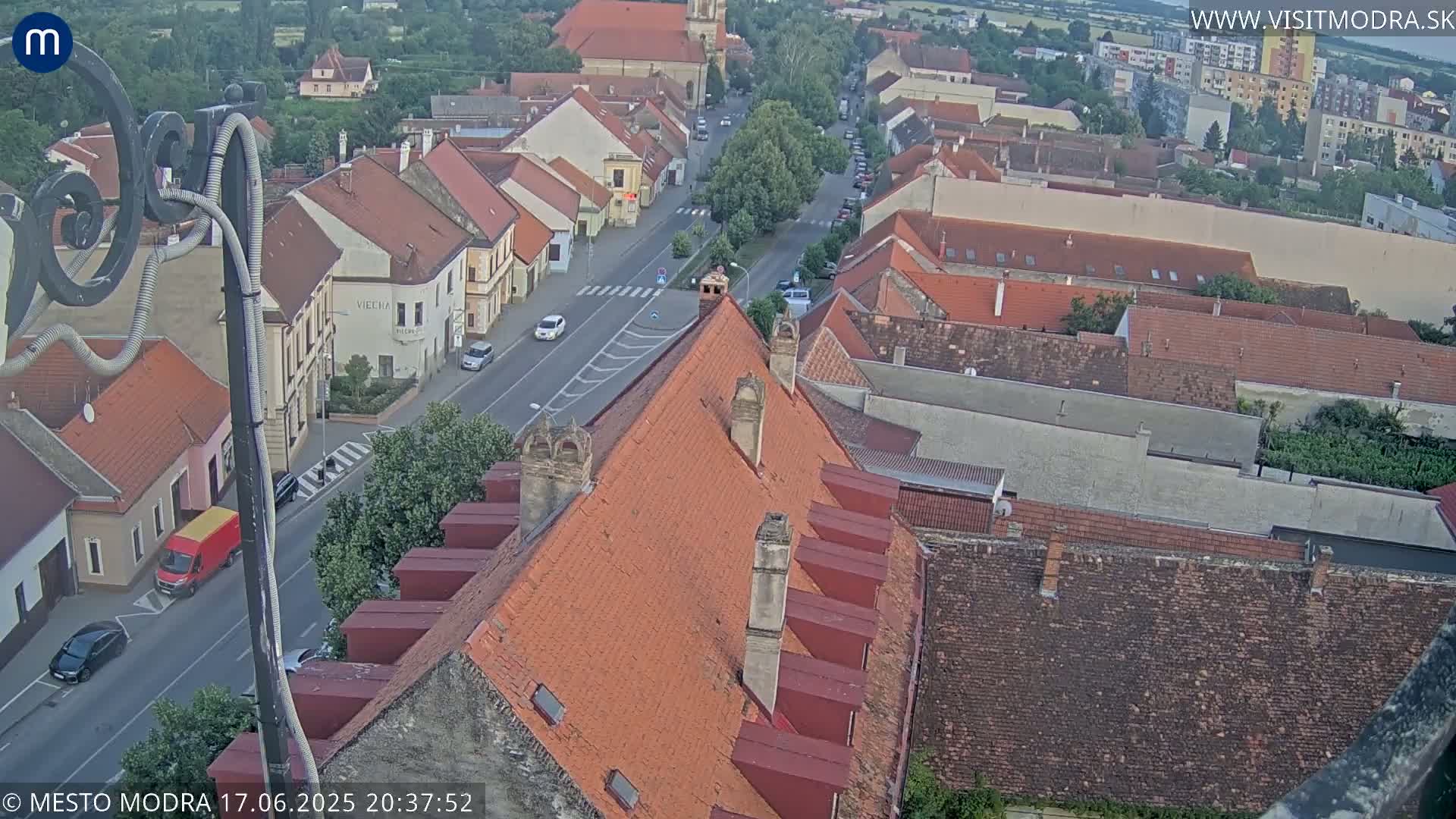 An aerial view of a town under a mostly clear evening sky shows red-tiled roofs of buildings lining a street with a few cars visible.
