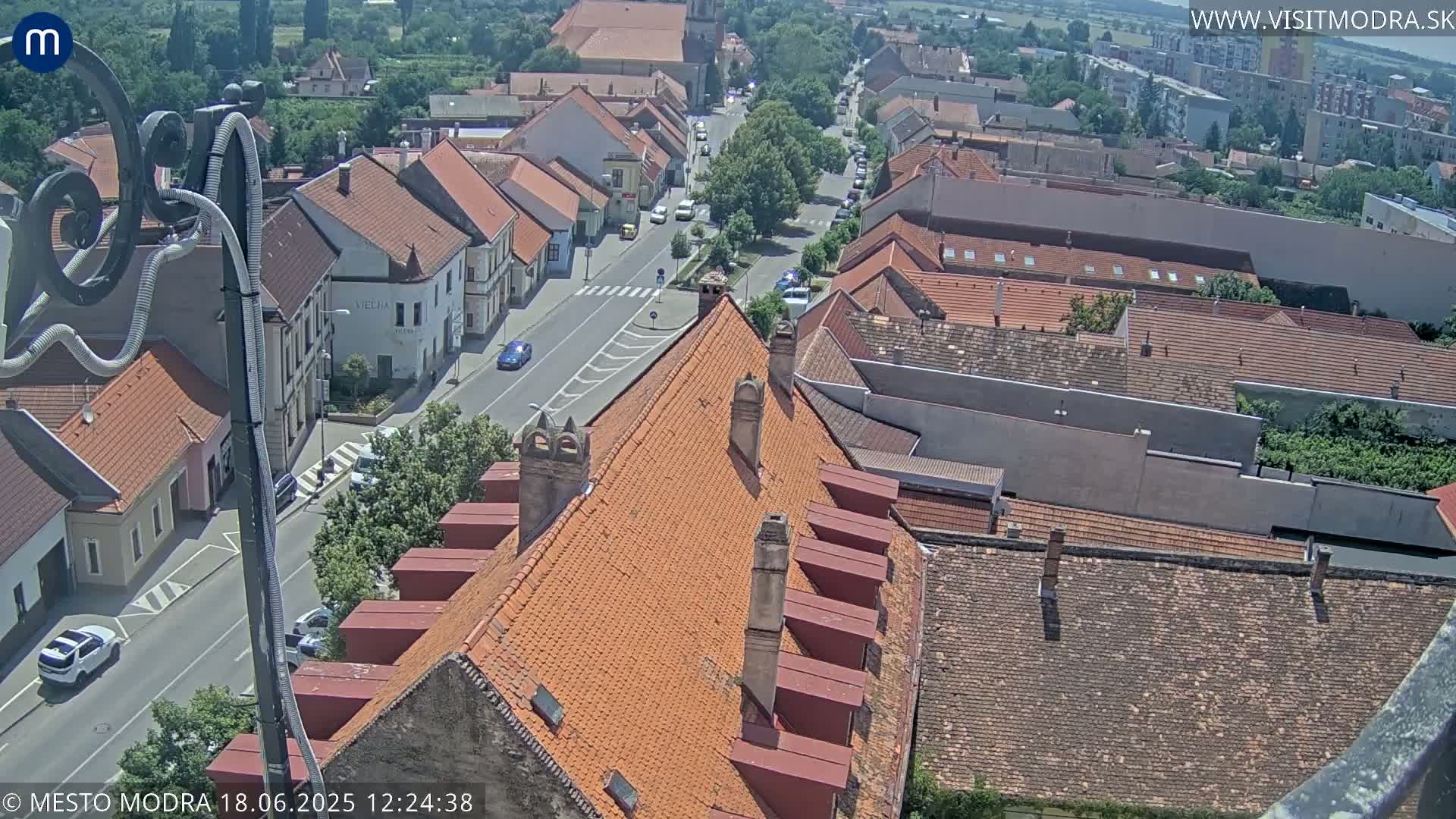 An aerial view of a town on a sunny day shows a street lined with red-tiled buildings, with cars parked along the road and other vehicles driving.