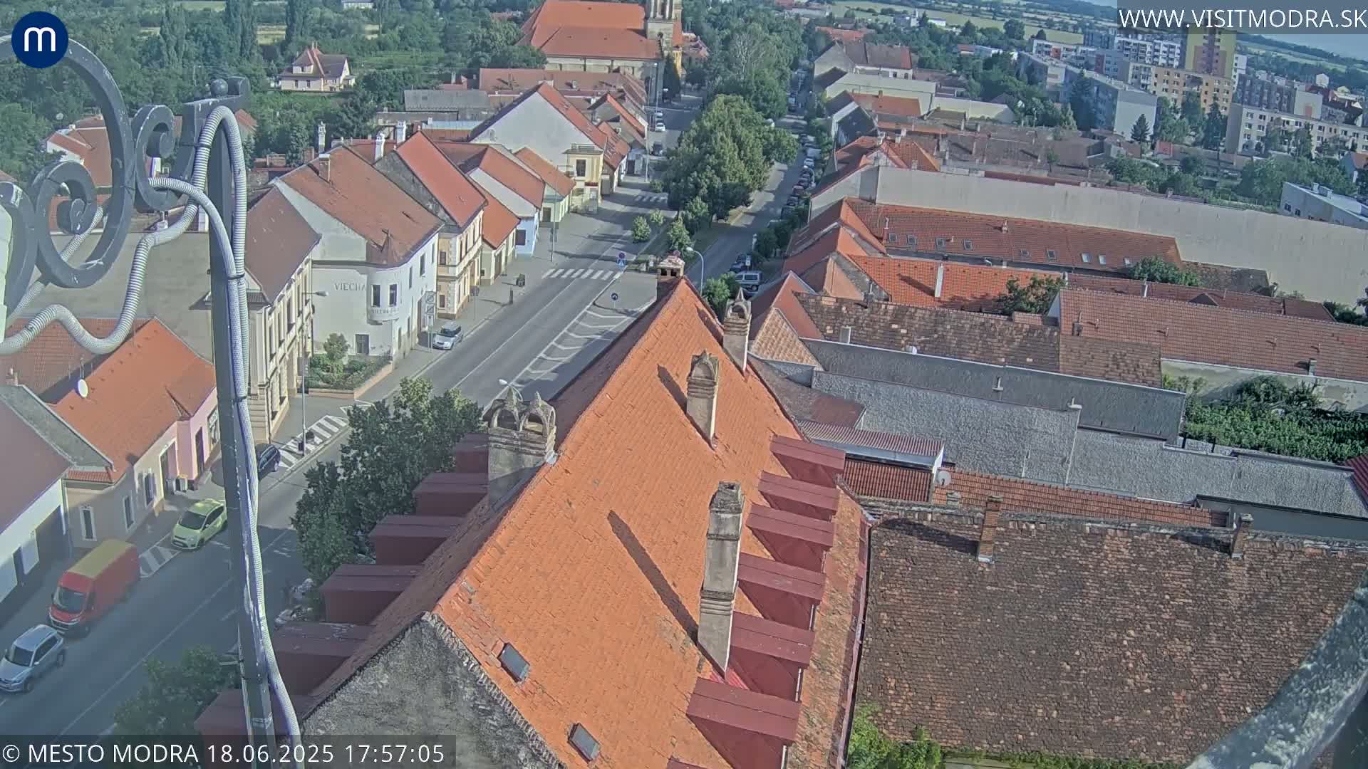 A high-angle, sunny view of a town's rooftops and streets, showing red tile roofs and a few cars.