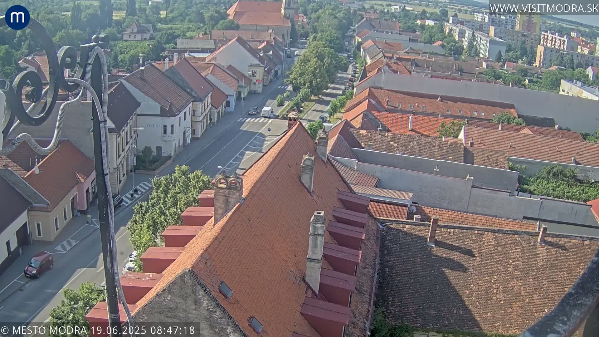 A high-angle view of a town with red tile roofs on sunny day, showing a street with cars and buildings stretching into the distance.