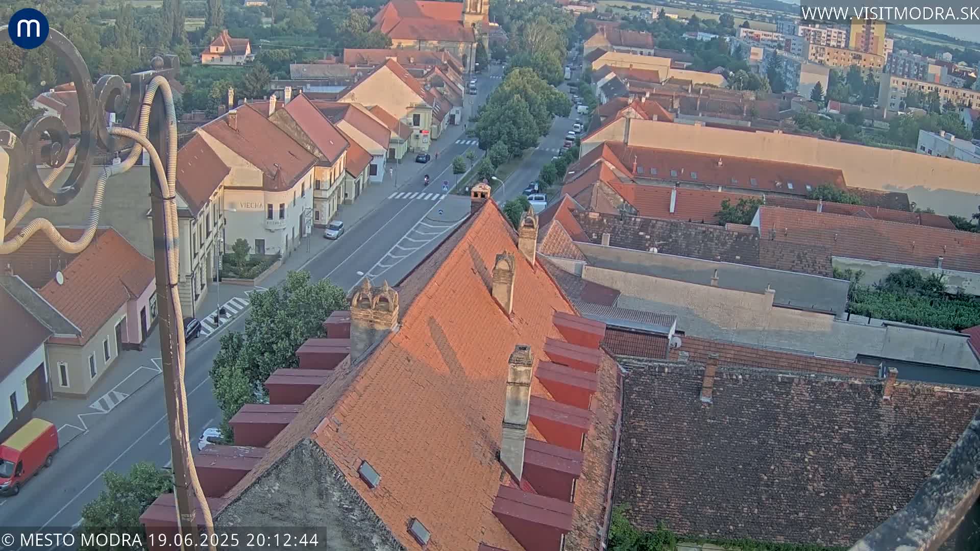 An aerial view of a town shows a street with cars and red-tiled roofs on buildings under a clear evening sky.