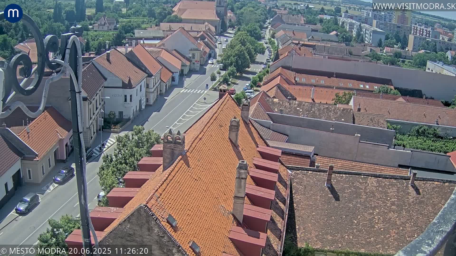 A high-angle view on a sunny day shows a town's streets and red-tiled roofs, with several cars visible on the roads.