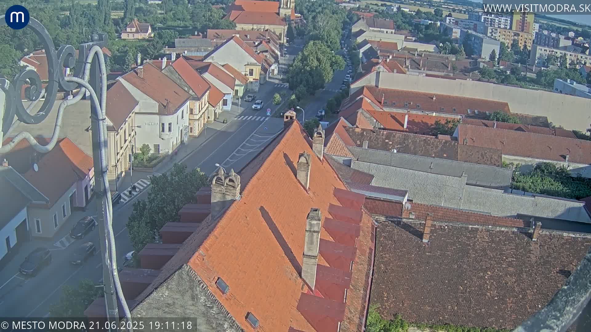 An aerial view of a town on a sunny day shows many red-tiled roofs and a street with cars.
