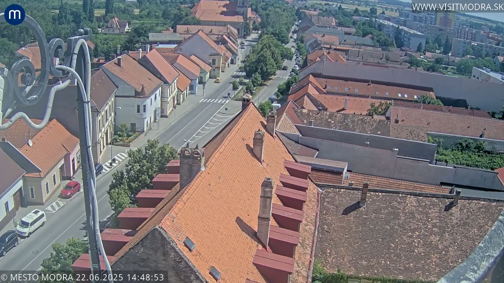 A high-angle view of a town on a sunny day, showing red-tiled rooftops, a street with cars, and surrounding buildings.