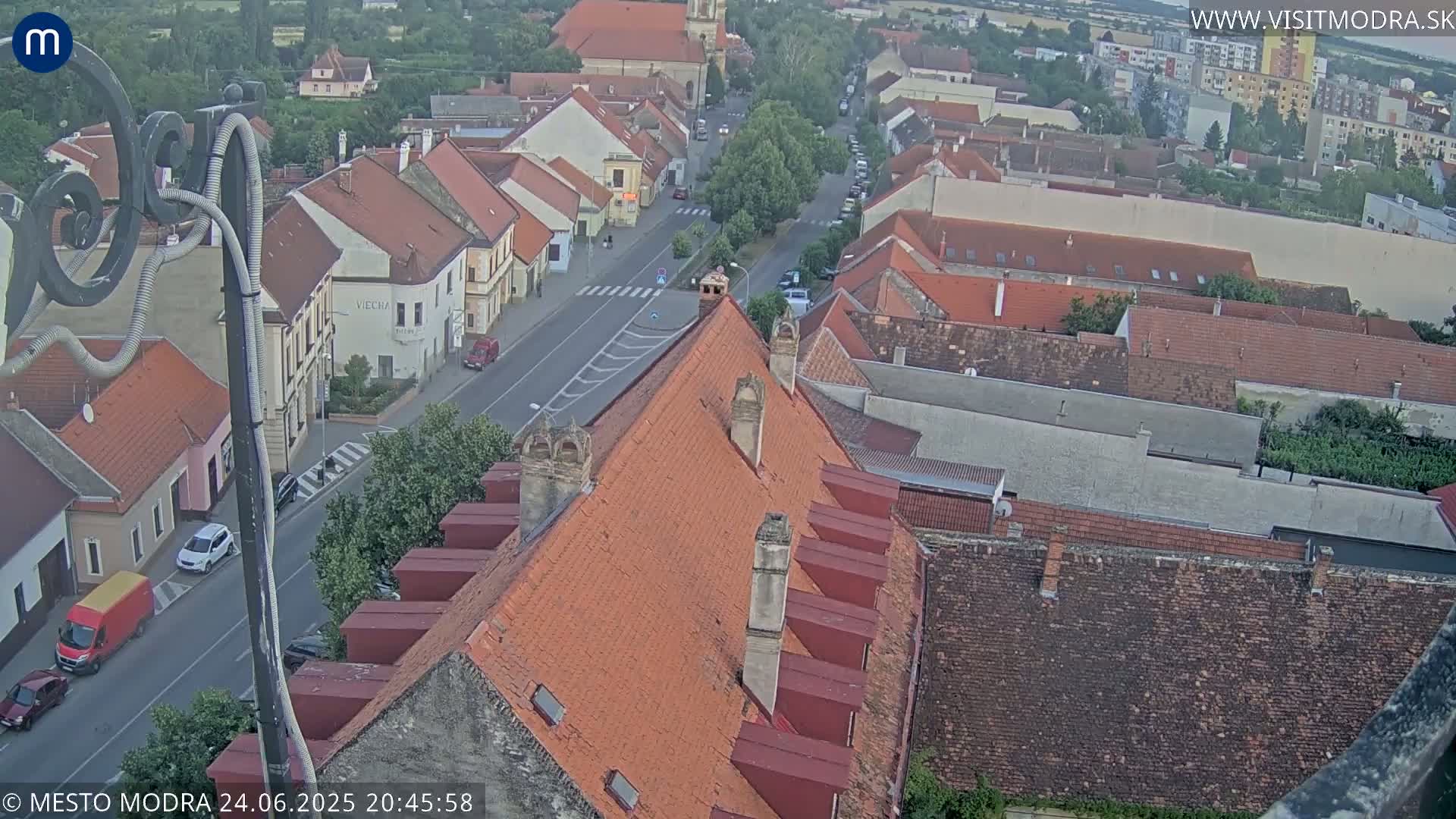 An aerial view of a town with red-tiled roofs and a street, under a partly cloudy sky.