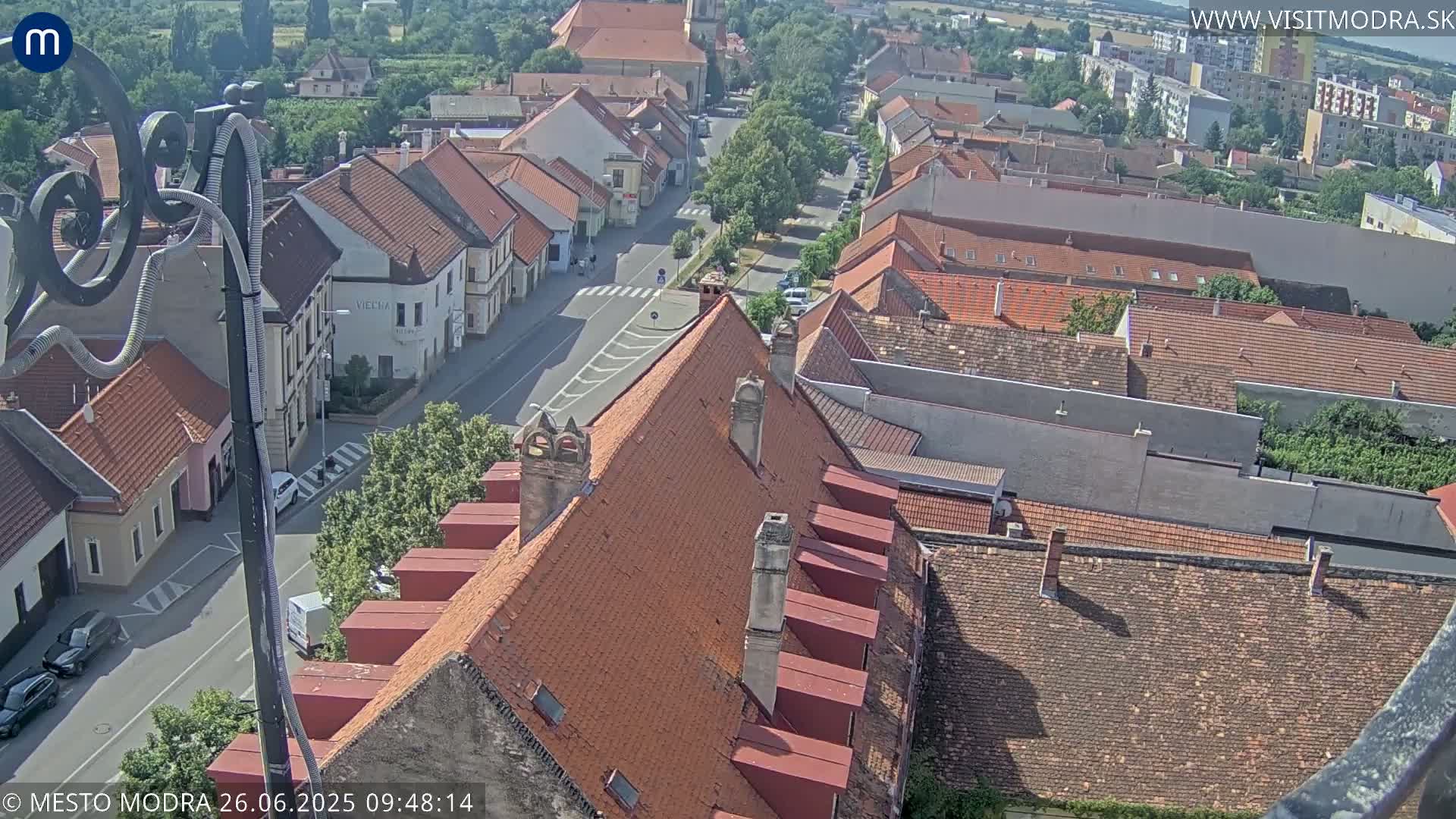 An aerial view of a town with red-tiled roofs and a street, under a partly cloudy sky.
