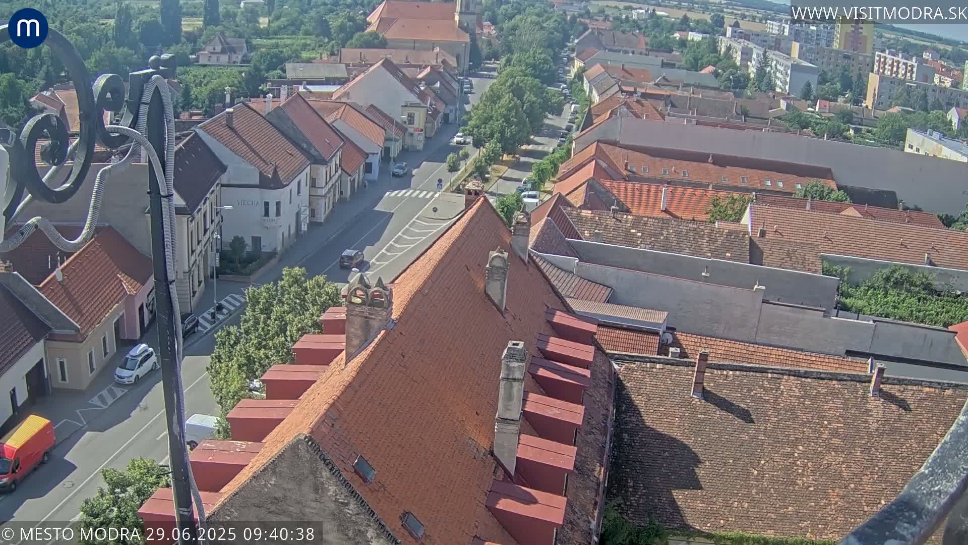 An aerial view of a town on a sunny day, showing a street lined with buildings and rooftops, with some vehicles visible.