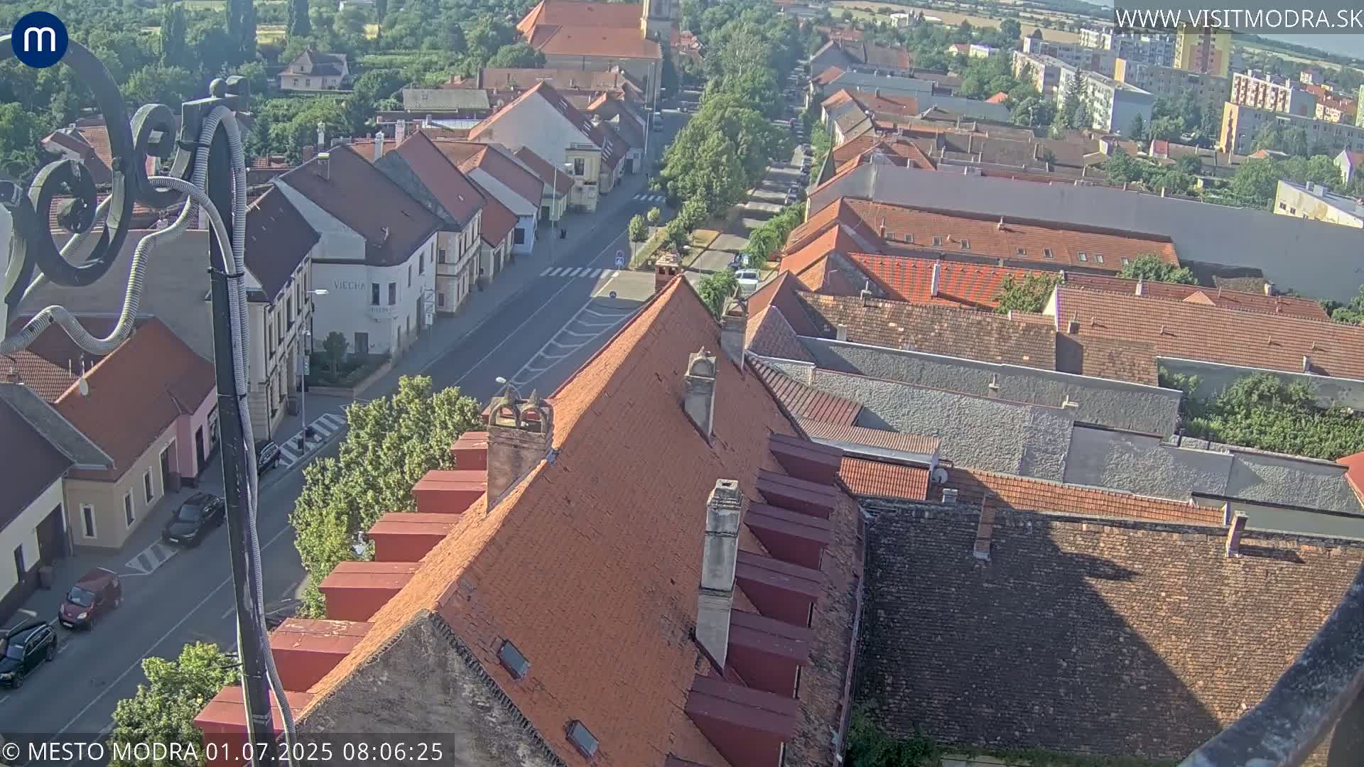 A high-angle view of a town on a sunny day shows numerous red-tiled roofs and a street with cars parked along the side.