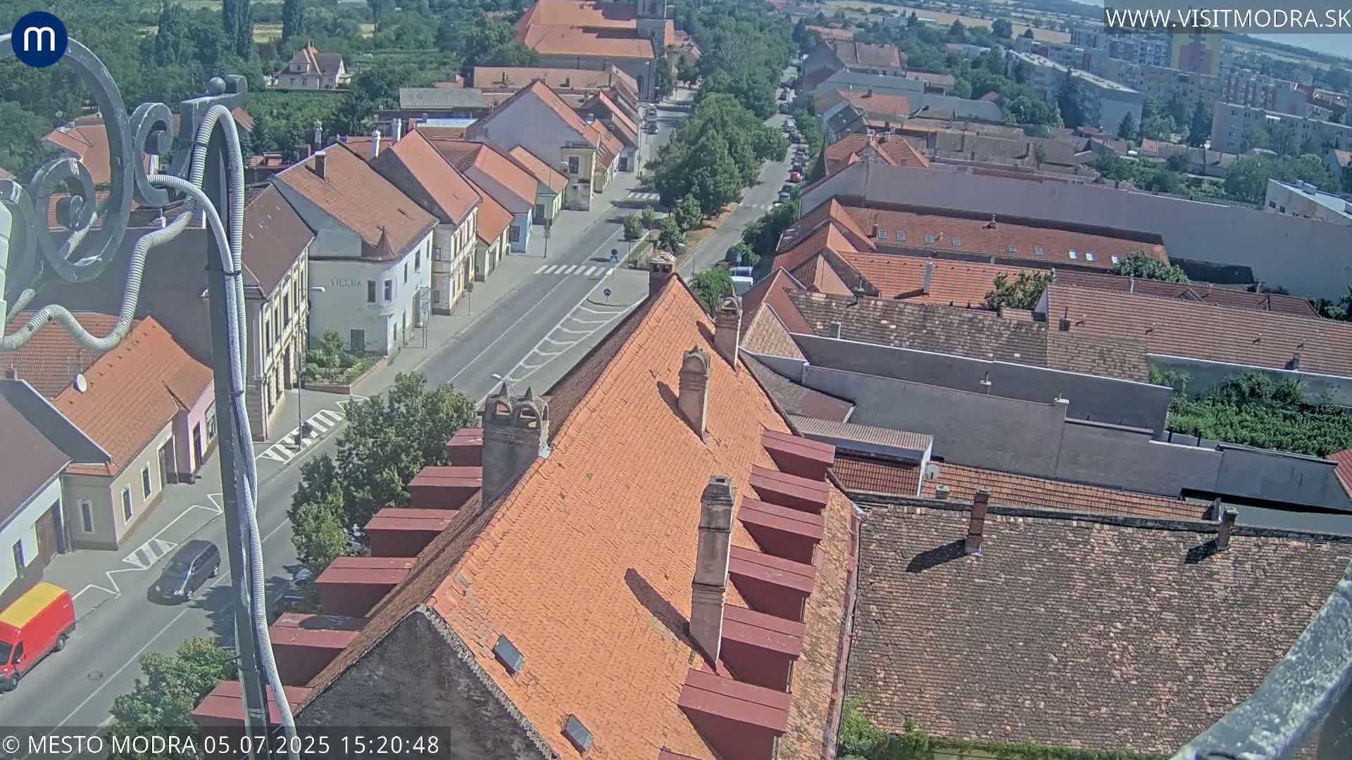 A high-angle, sunny view of a town's red-tiled rooftops and streets, with a few vehicles visible.