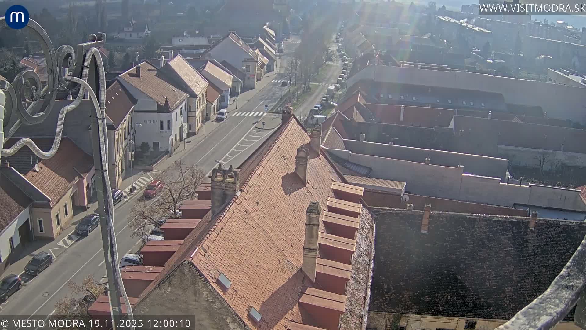 A high-angle, sunny view shows a European town with red-tiled roofs, a church steeple, and a bird perched on a building in the foreground.
