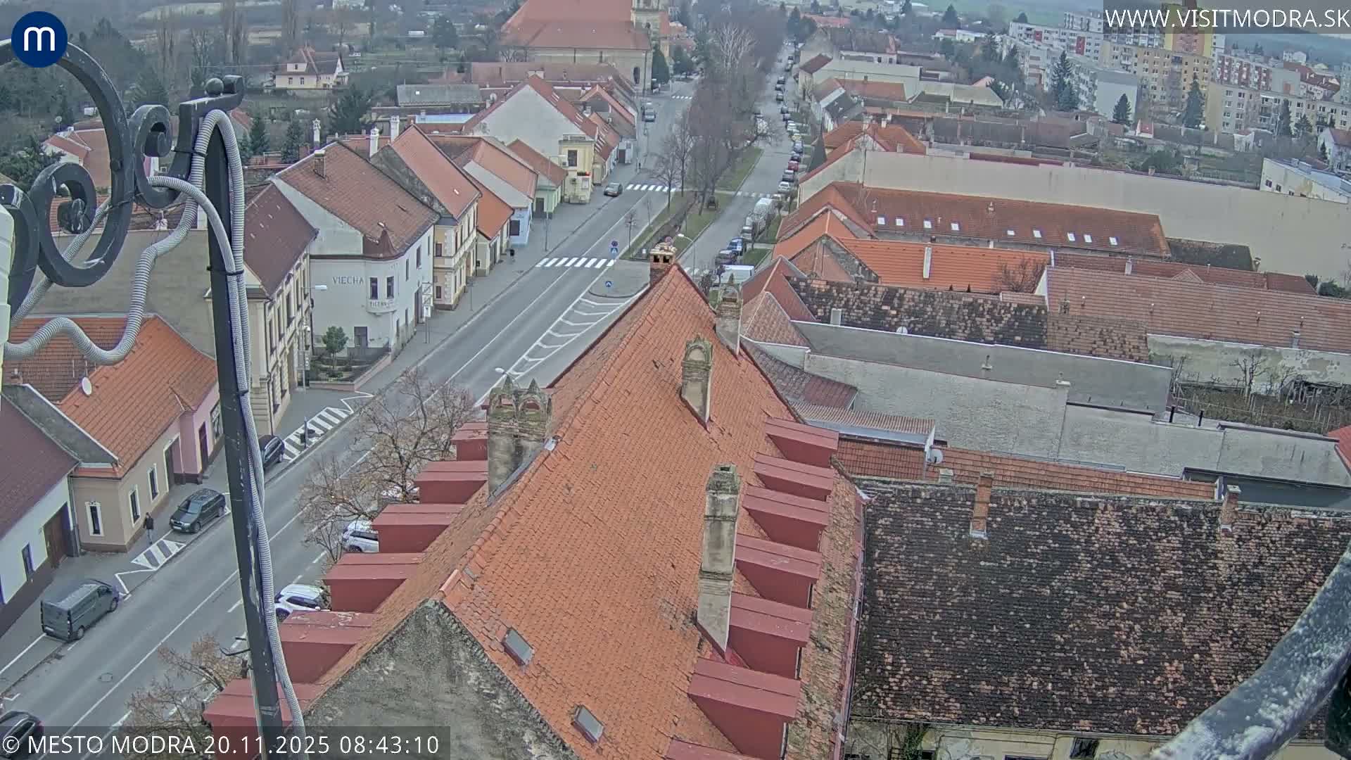 An elevated view reveals a European town's street scene with numerous buildings sporting red-tiled roofs, parked vehicles, and a few pedestrians, all under an overcast sky.