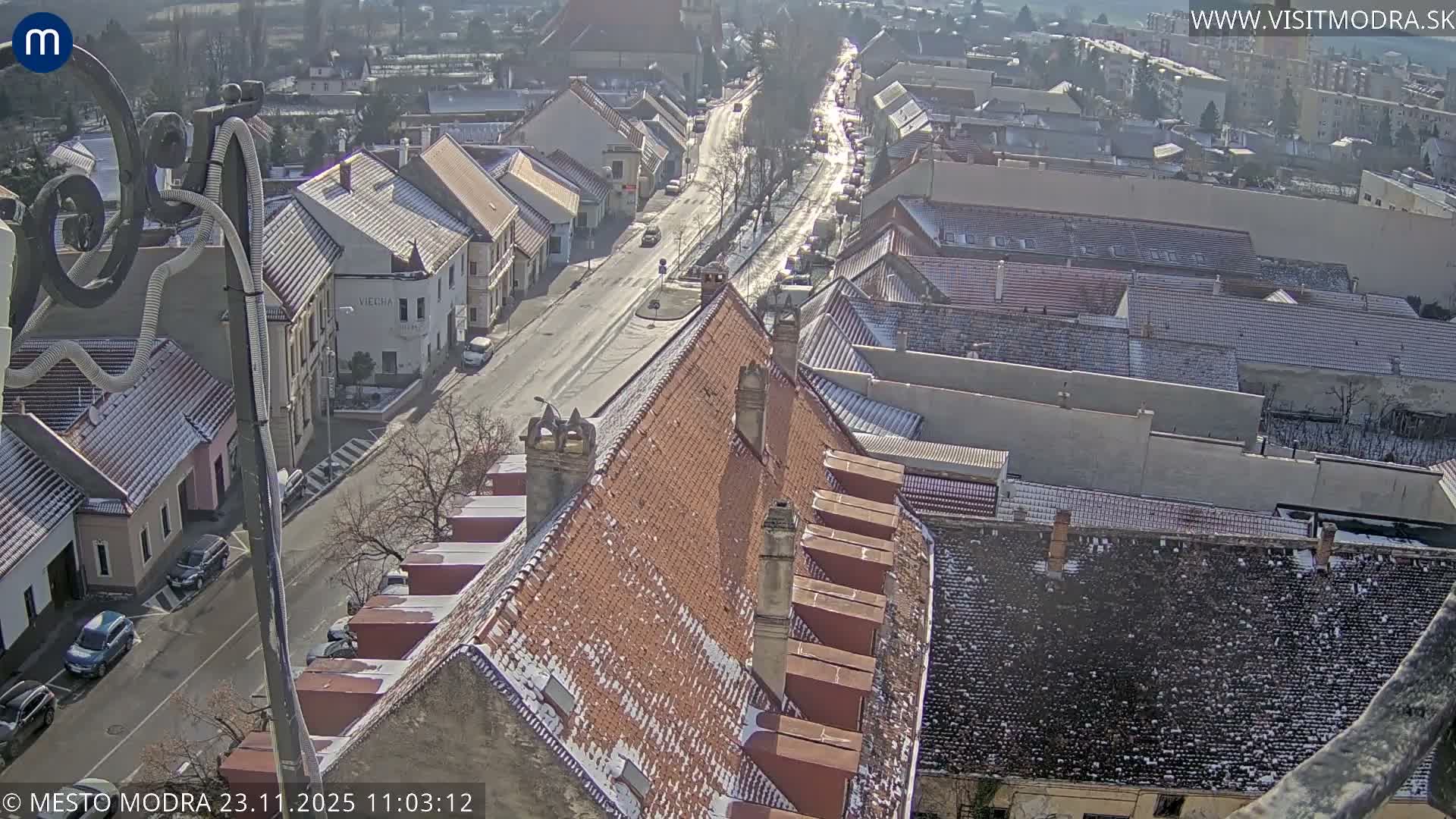 The image shows a clear and sunny winter day overlooking a European town, with a light dusting of snow visible on the rooftops of the buildings and bare trees lining the streets.