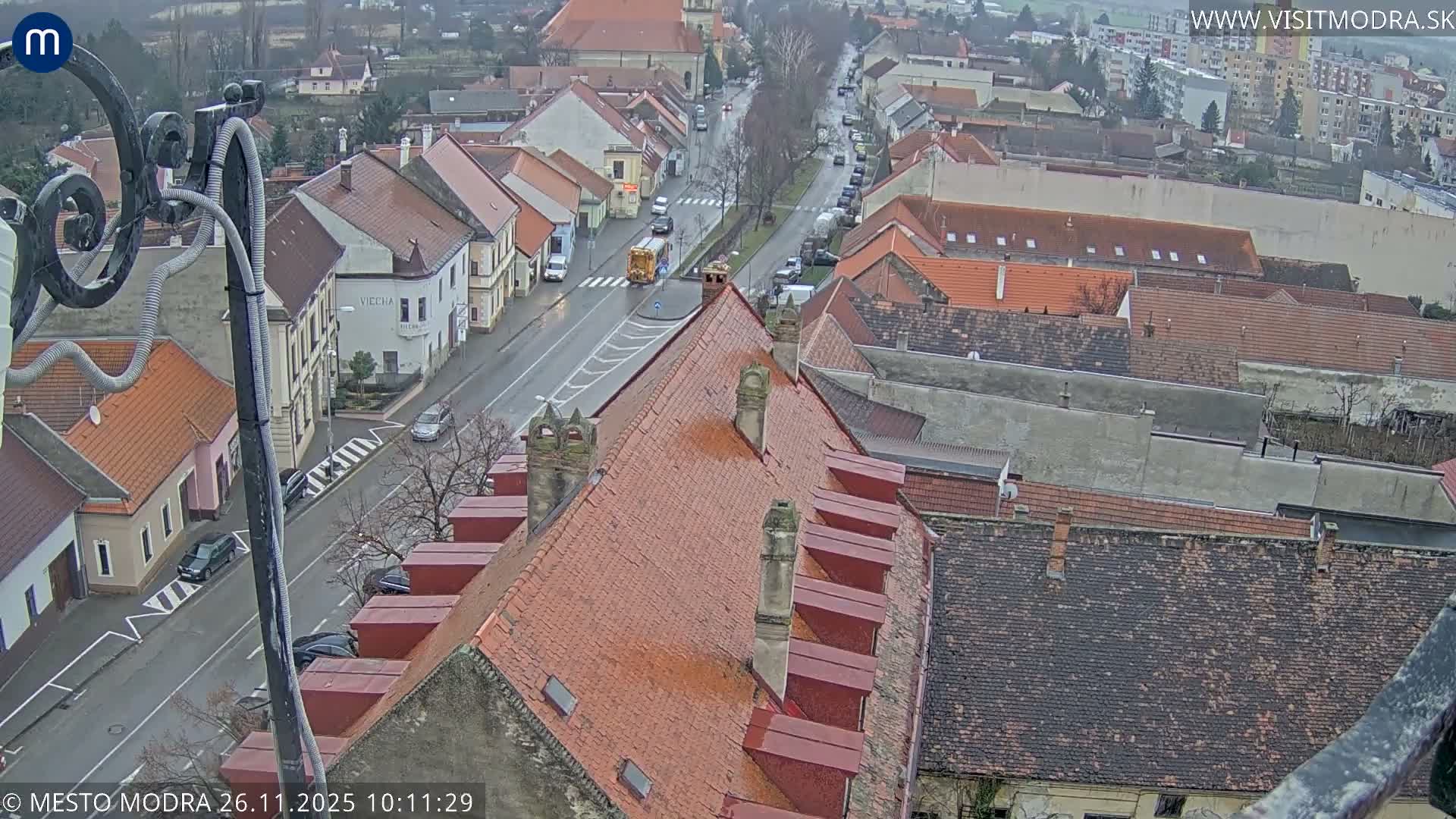 From an elevated perspective, a wide red-tiled roof dominates the foreground of a damp European town street bustling with cars and a yellow bus under an overcast sky, extending to residential buildings in the distance.