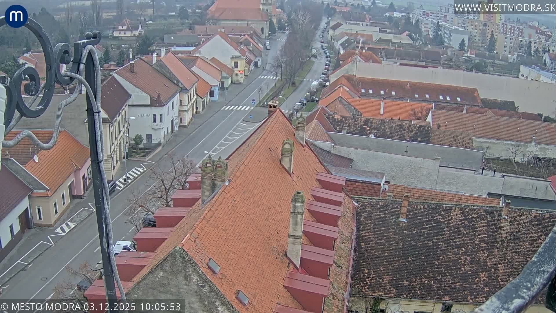 An overcast, high-angle view shows a European town street, partially framed by an ornate metal structure, flanked by buildings with red-tiled roofs and bare trees, with scattered cars, all receding into a hazy background under a uniform gray sky.
