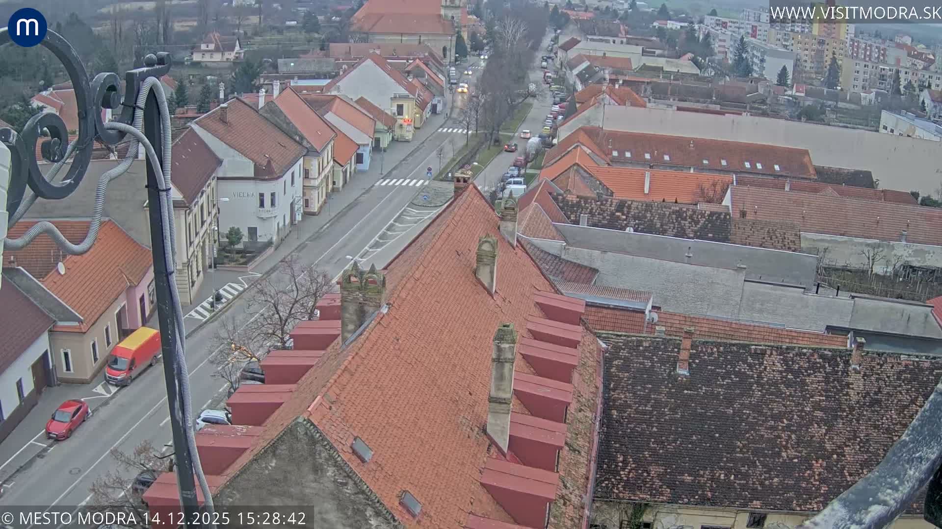 An overcast, high-angle view shows a European town street, partially framed by an ornate metal structure, flanked by buildings with red-tiled roofs and bare trees, with scattered cars, all receding into a hazy background under a uniform gray sky.