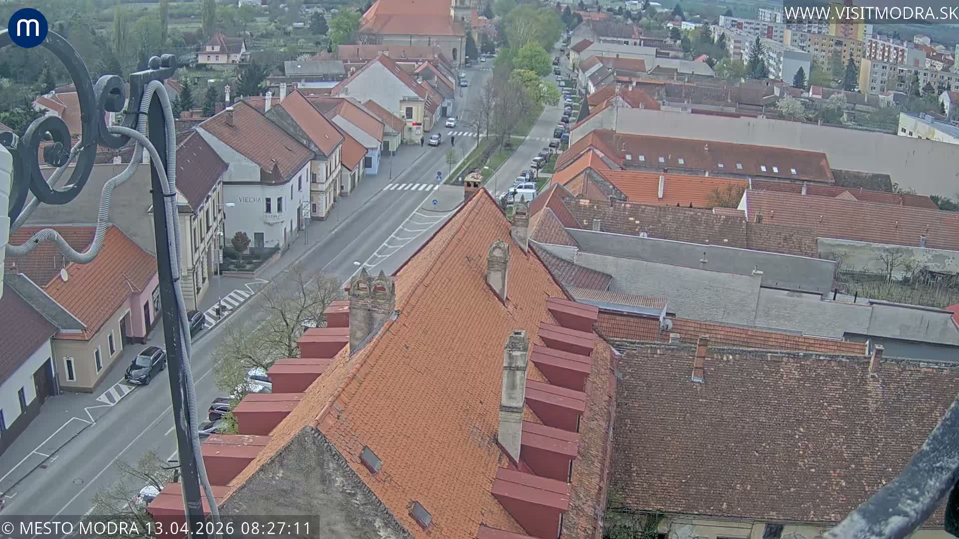 An overcast, high-angle view shows a European town street, partially framed by an ornate metal structure, flanked by buildings with red-tiled roofs and bare trees, with scattered cars, all receding into a hazy background under a uniform gray sky.