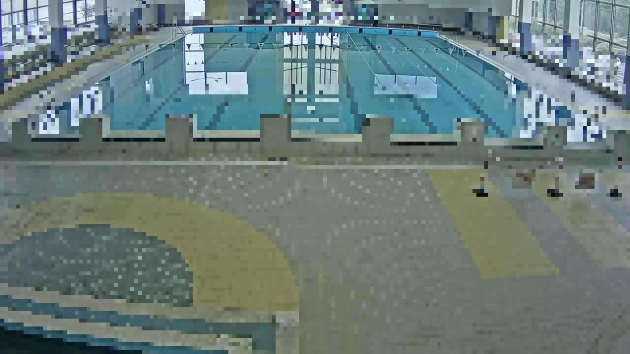 An indoor swimming pool with several lanes is visible from an elevated vantage point, the surrounding floor is mainly gray tile with a section of tan tile.