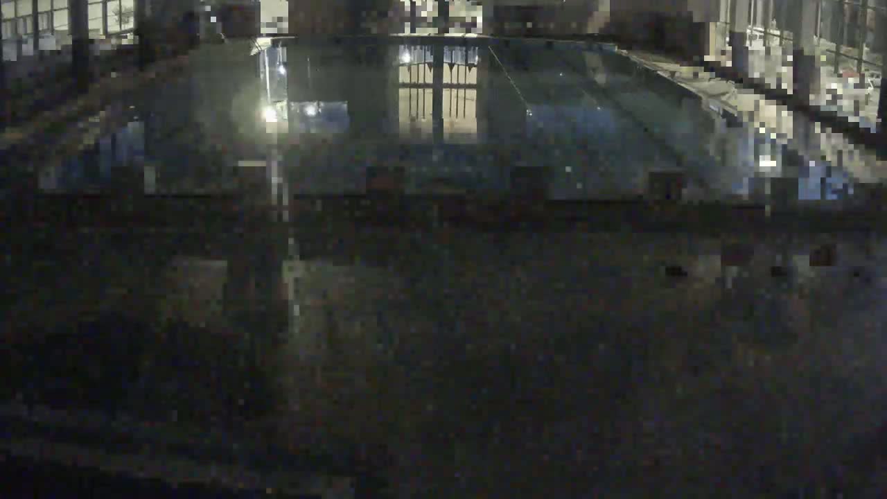 A nighttime, overhead view shows a dark, seemingly empty indoor swimming pool reflecting dim lights and a building's interior.