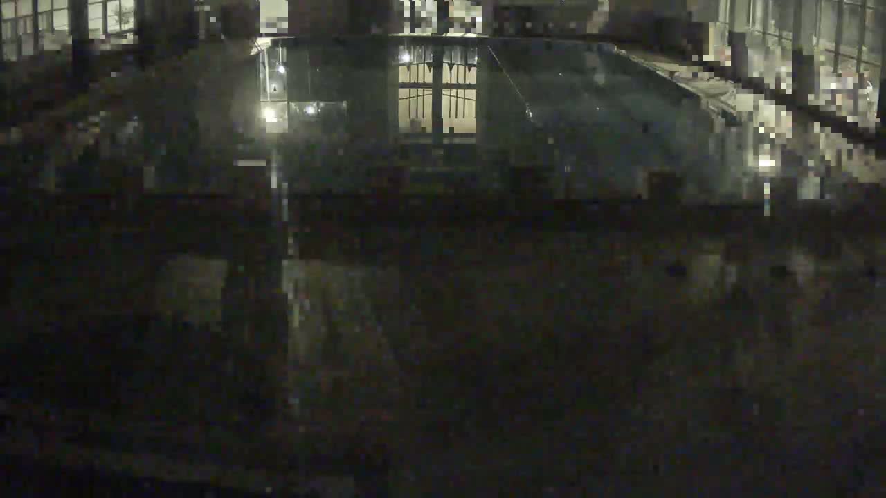 A dark, indoor swimming pool is seen at night, reflecting the lights and windows of the building.