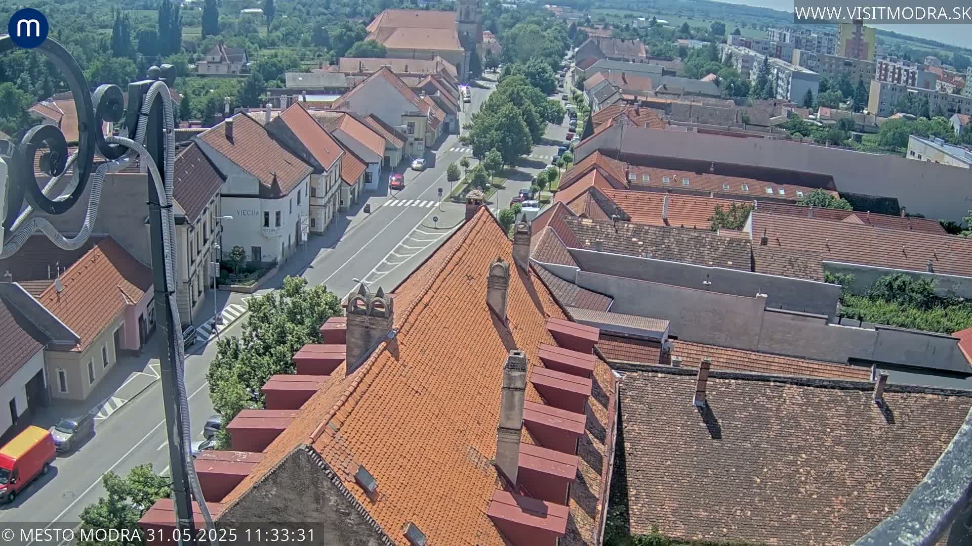 An aerial view on a sunny day shows a town square with several buildings and a street, some with orange-tiled roofs.